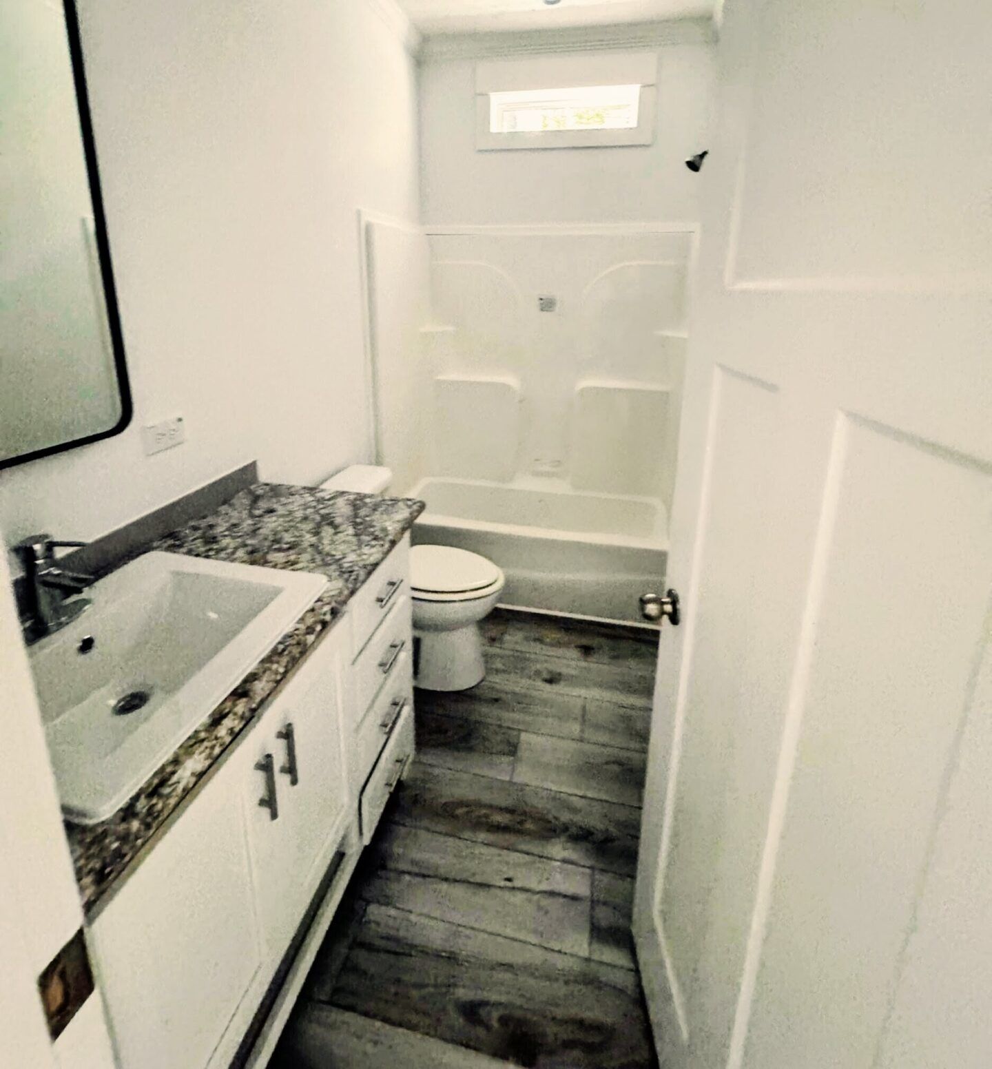 Small white bathroom with granite vanity, toilet, tub-shower, and narrow gray wood-look floor.