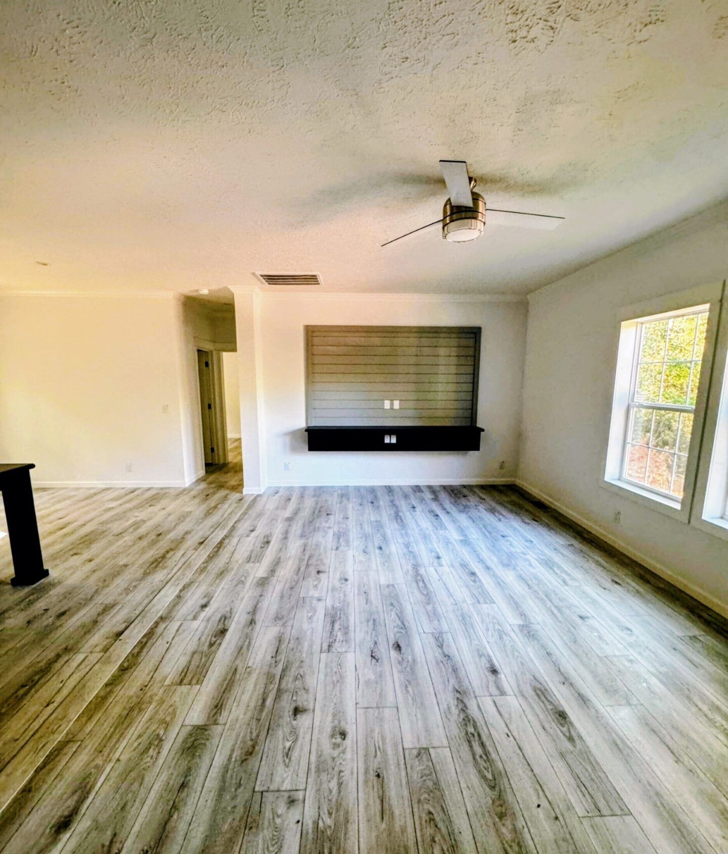 Empty modern living room with light wood floors, white walls, ceiling fan, and large windows.