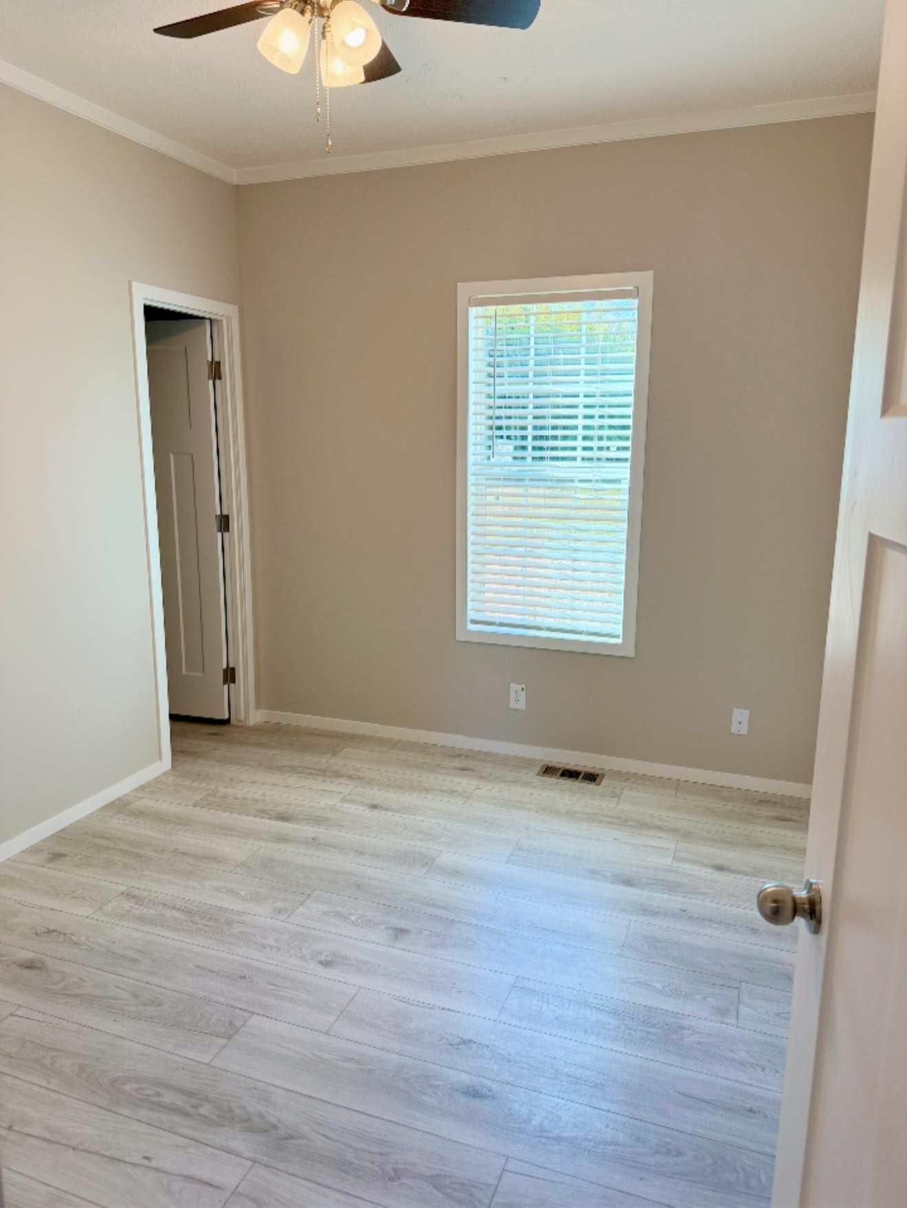 Empty beige bedroom with white trim, window blinds, light wood floor, and ceiling fan.