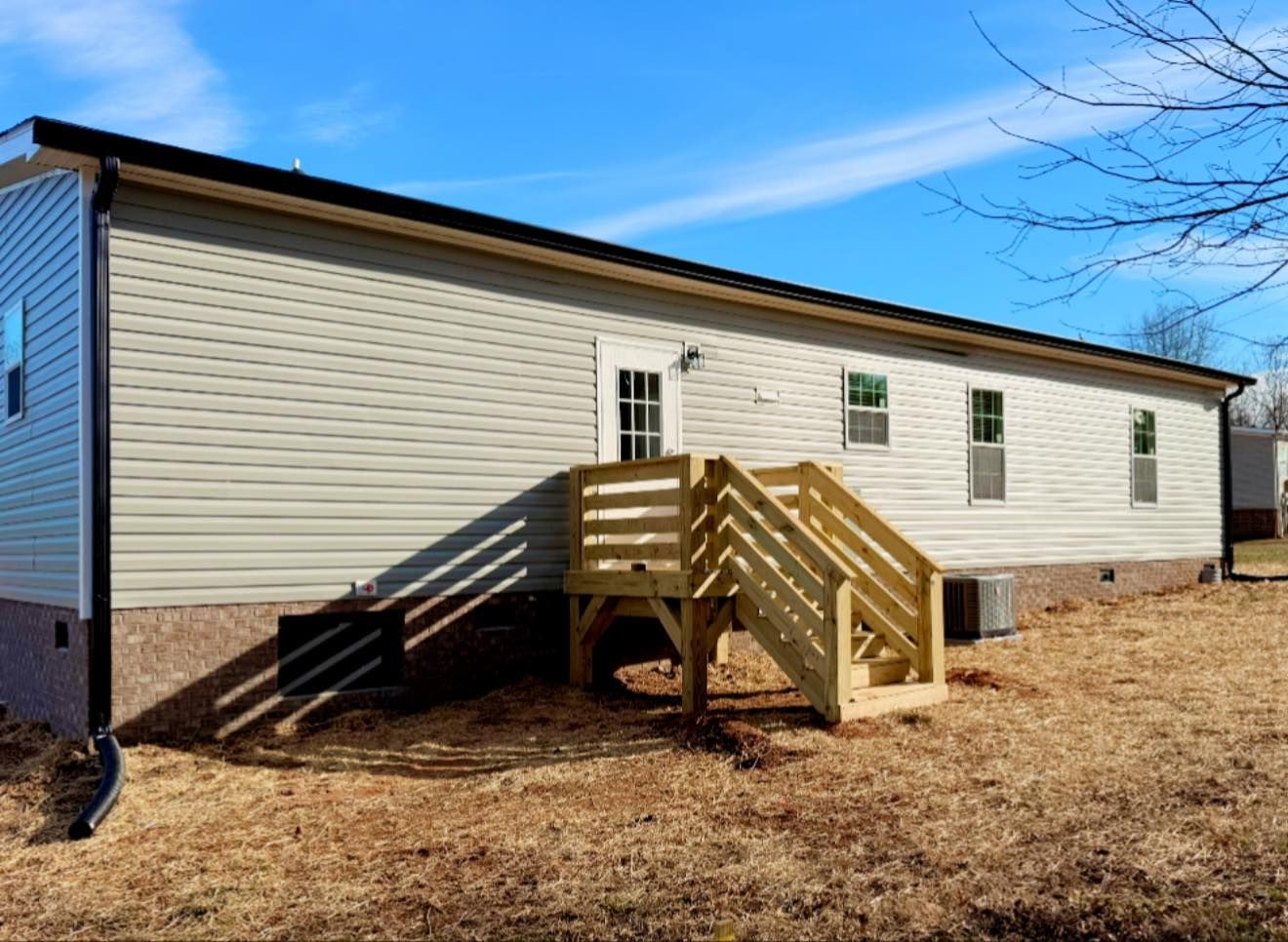 Single-story beige house with a wooden front deck and steps on a dry yard under a clear blue sky