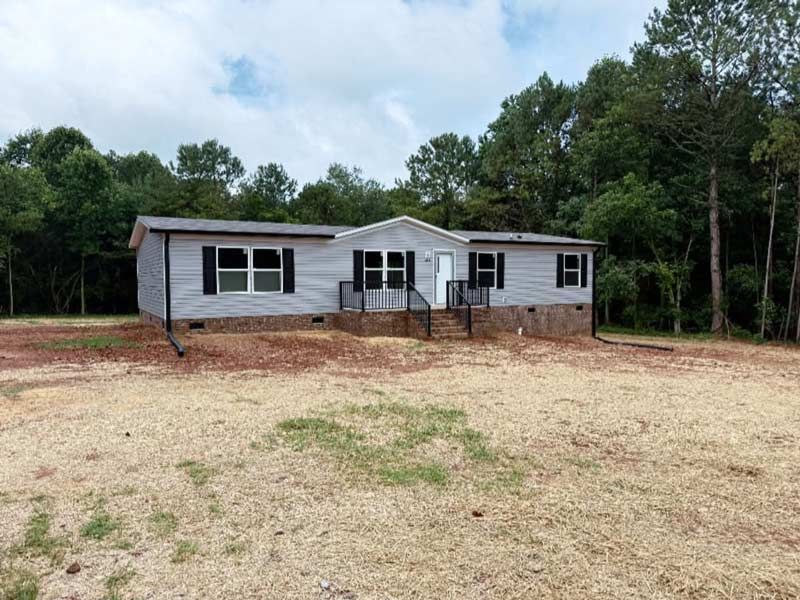 Single-story gray house with a front porch, set on a bare lot surrounded by trees.