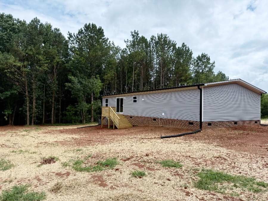 Single-story white house with a yellow porch on a dirt lot beside a wooded area
