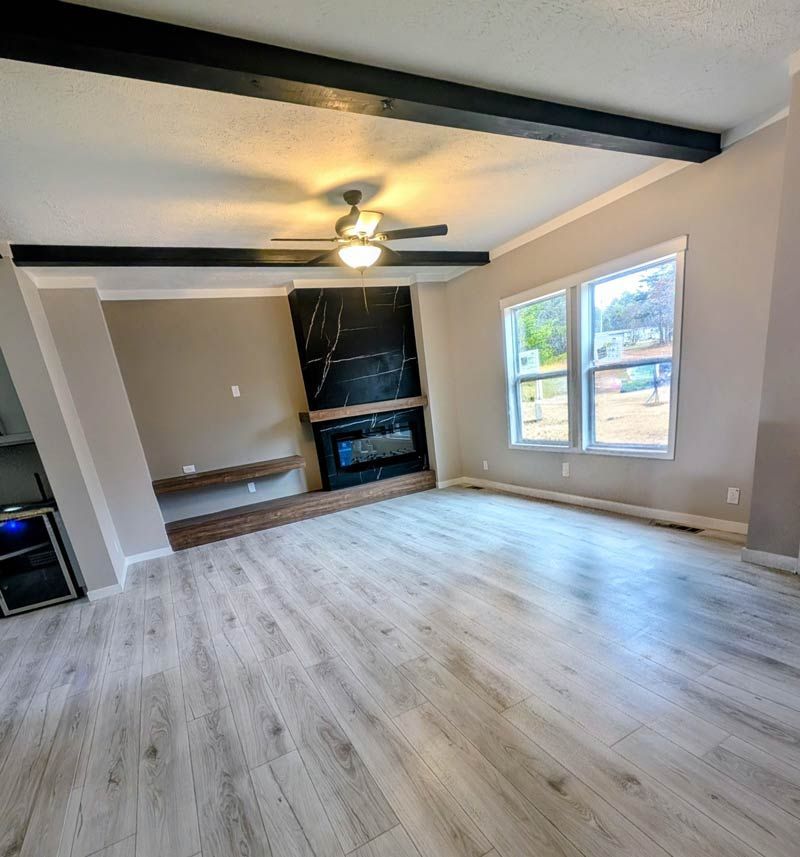 Empty living room with light wood floors, black ceiling beams, a fireplace, and large windows.