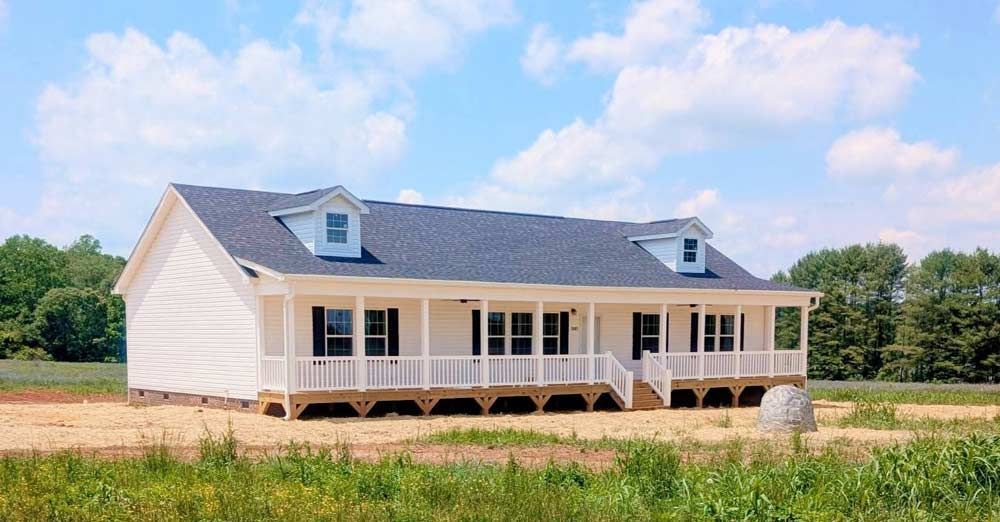 New white farmhouse with porch and gray roof in a grassy field under a blue sky