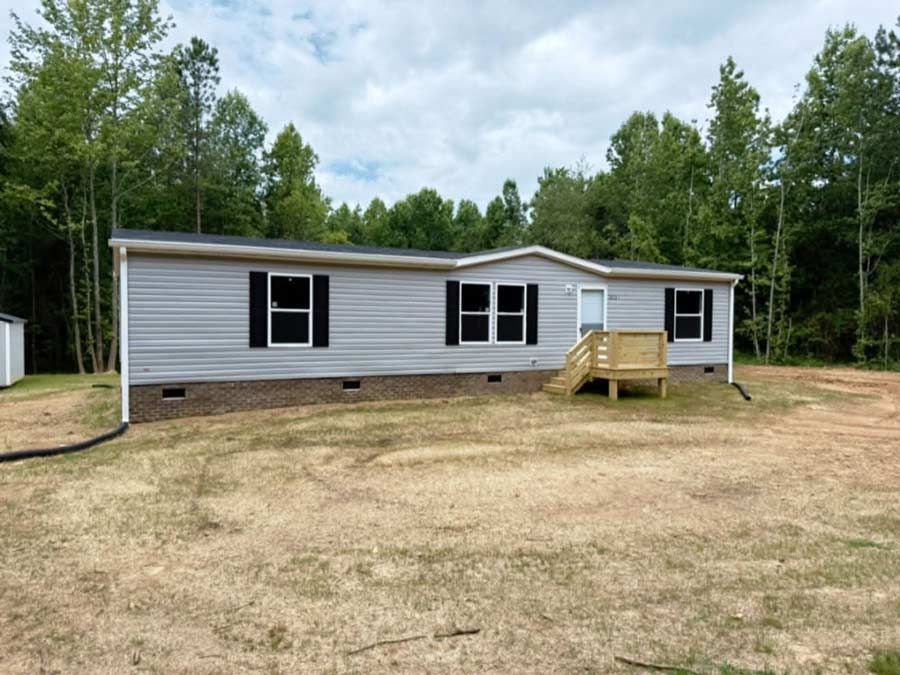 Single-story gray manufactured home with a small wooden porch, set in a grassy clearing by trees.
