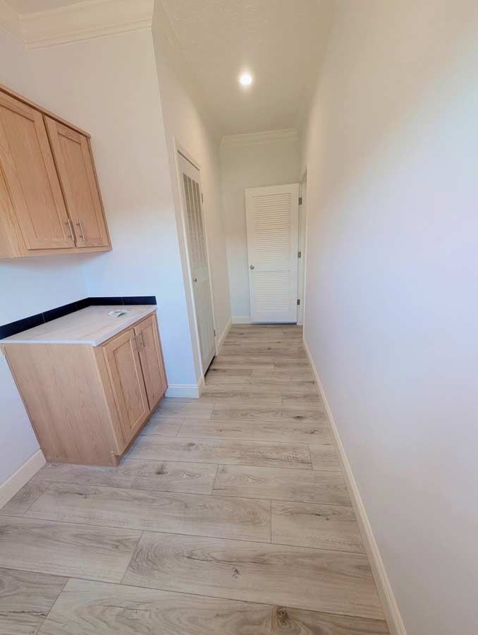 Narrow hallway with light wood cabinets, white walls, and pale wood flooring leading to a closed door.