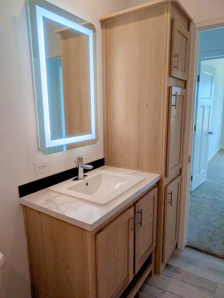 Bathroom vanity with light wood cabinets, white sink, black countertop, and lit mirror in a hallway.