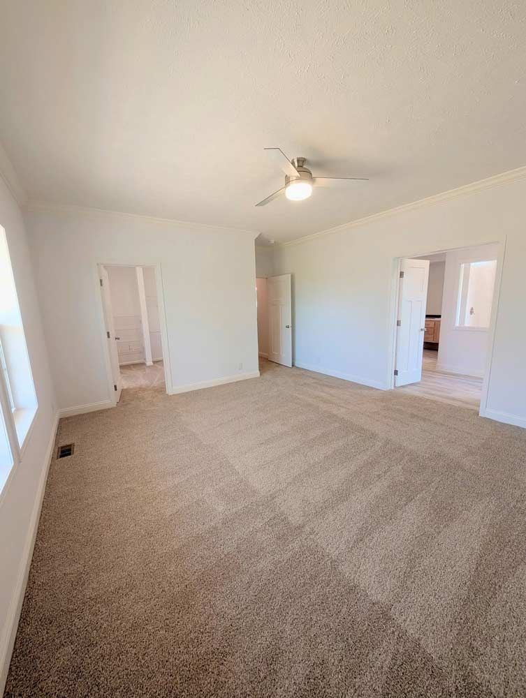 Empty beige-carpeted bedroom with white walls, ceiling fan, and open doorways to adjacent rooms