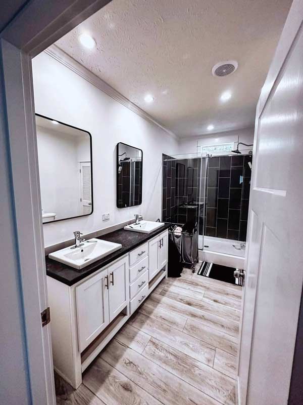 Narrow modern bathroom with double vanity, mirrors, wood-look floor, and glass-enclosed shower.