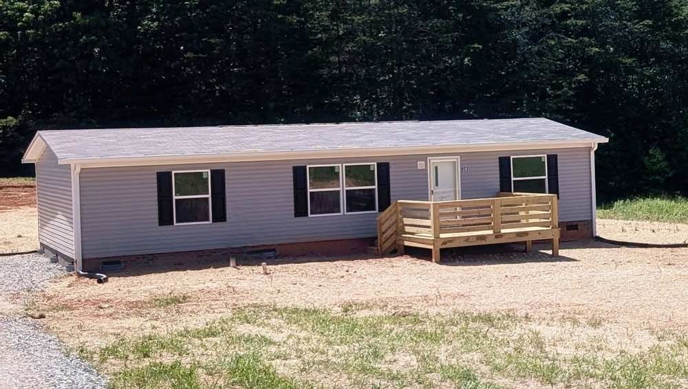 Single-story beige mobile home with a small wooden porch in a wooded lot