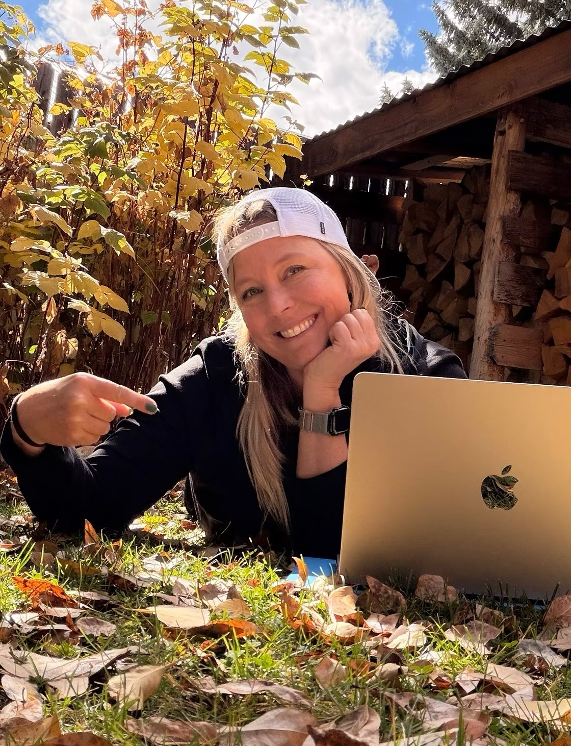 Leia Morrison lying on autumn leaves beside a laptop near stacked firewood under golden trees