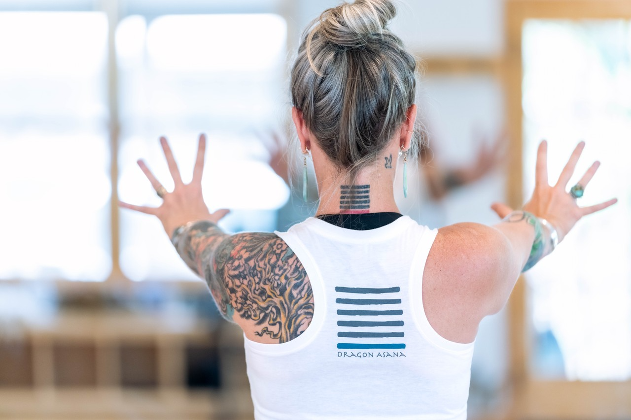 Person with tattooed back raises hands in a yoga studio with mirrors and large windows.