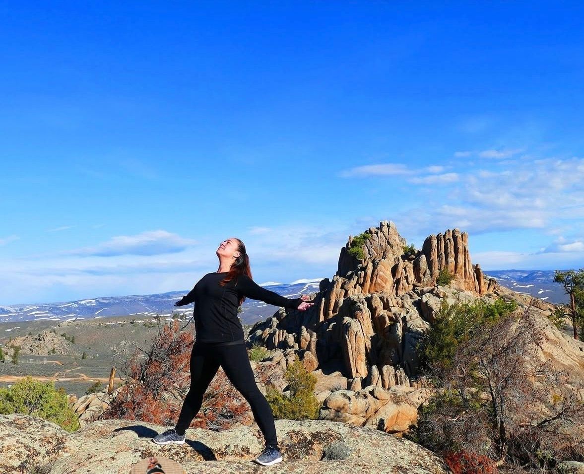 Person in black clothes posing on a rocky overlook under a bright blue sky, with a jagged mountain behind.
