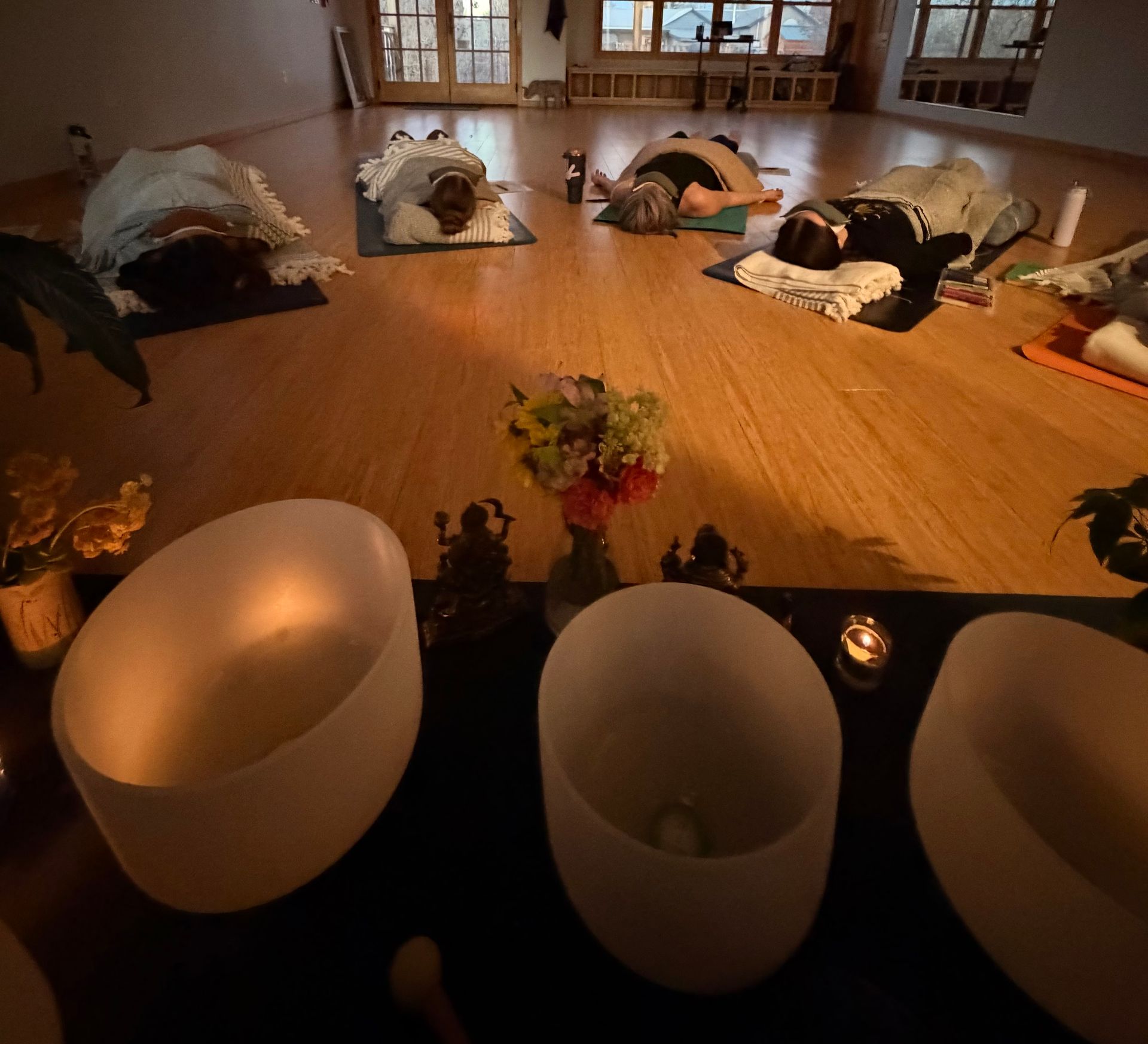 Dimly lit meditation room with people lying on mats, surrounded by singing bowls and candles.