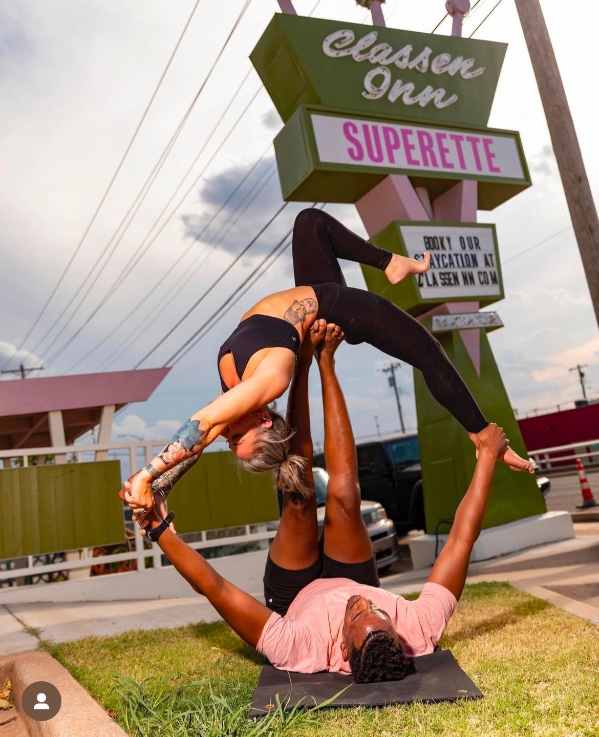 Two people doing a partner yoga pose outdoors beside a tall “Superette” sign.