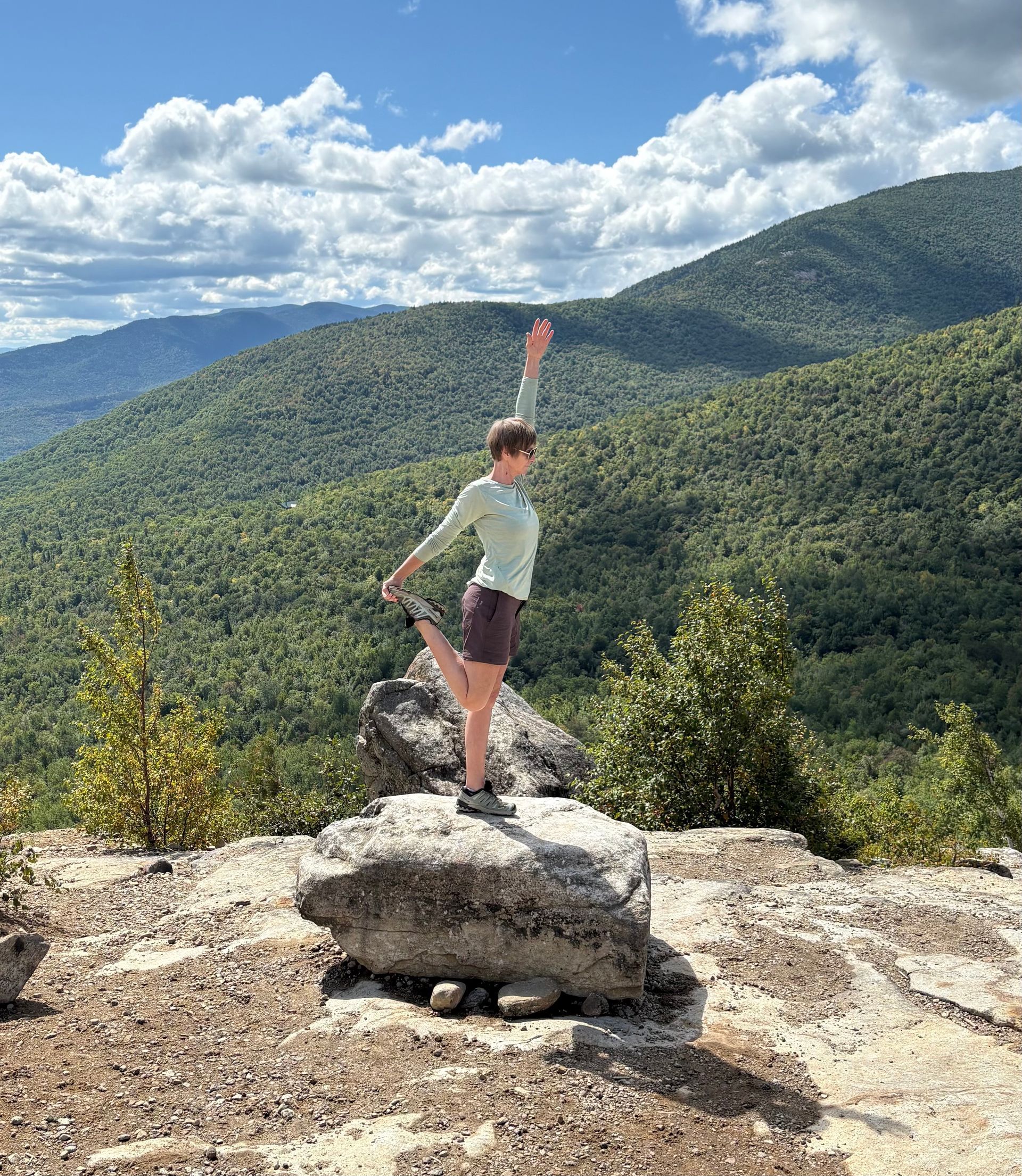 Person standing on a rocky overlook with one arm raised, overlooking forested mountains under a cloudy sky