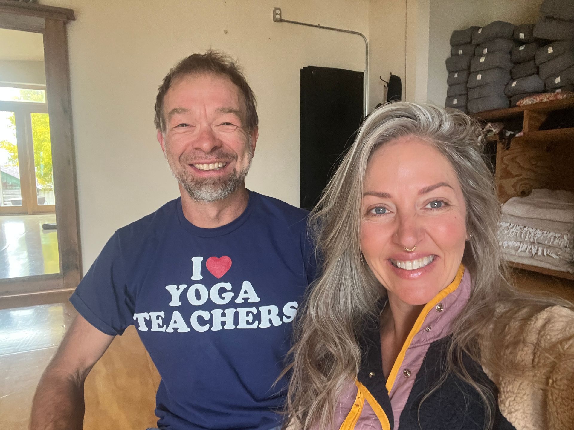 Two smiling people standing indoors, one wearing an “I ❤️ YOGA TEACHERS” shirt.