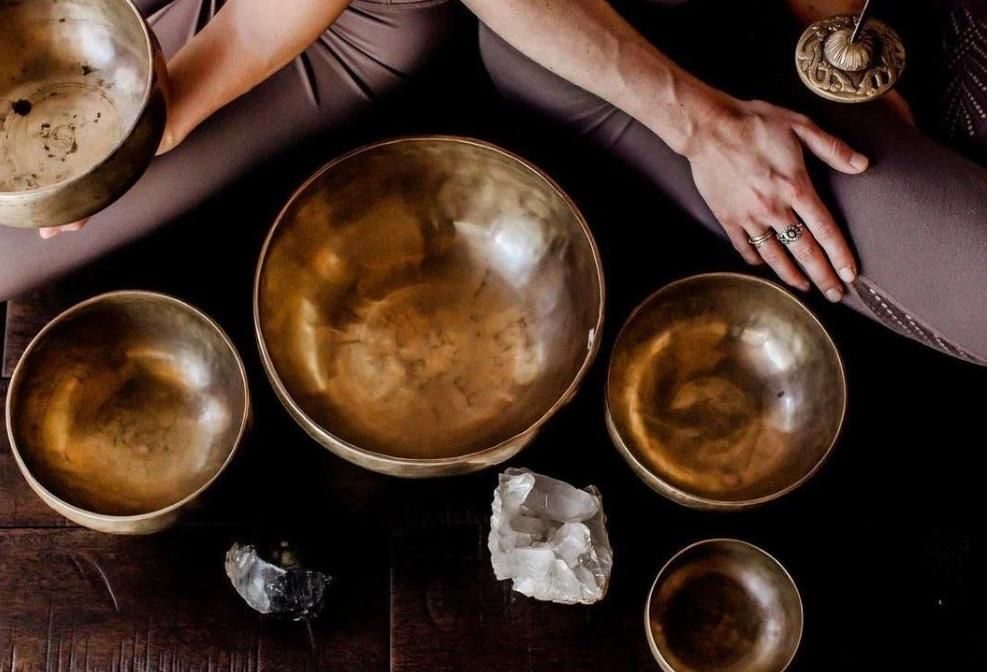 Assorted brass singing bowls with hands resting on cushions and a crystal on a dark surface