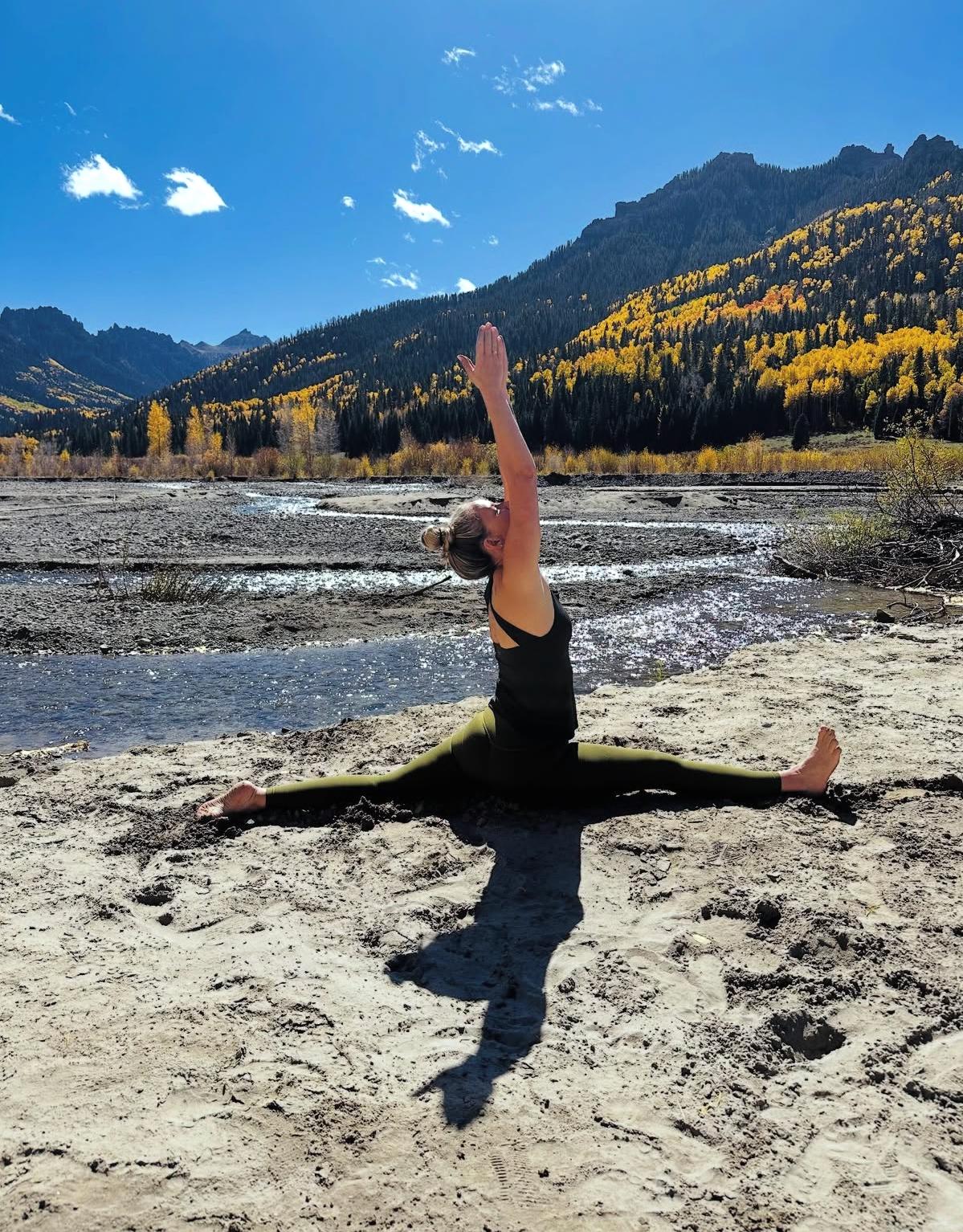 Person doing a split on a sandy riverbank with mountains and autumn trees in the background.