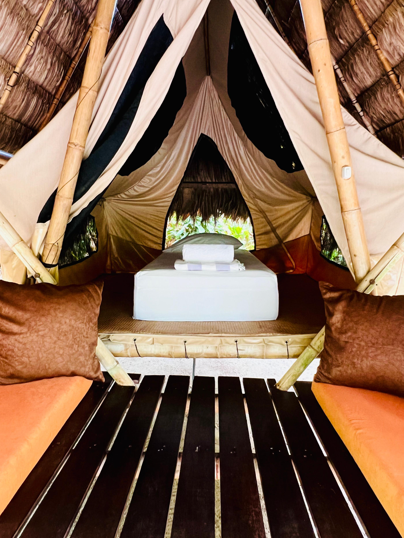Inside a canvas tent, a low bed with white linens and brown pillows faces an opening to greenery.