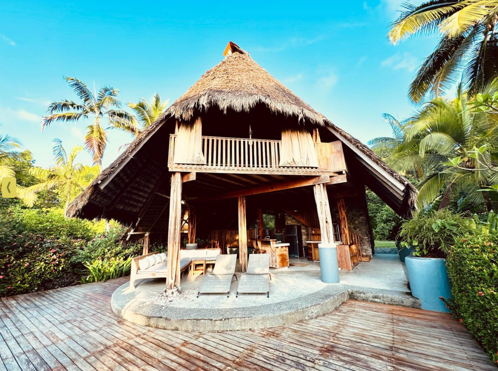Thatched tropical beach hut with wooden deck, lounge chairs, and palm trees under a bright blue sky