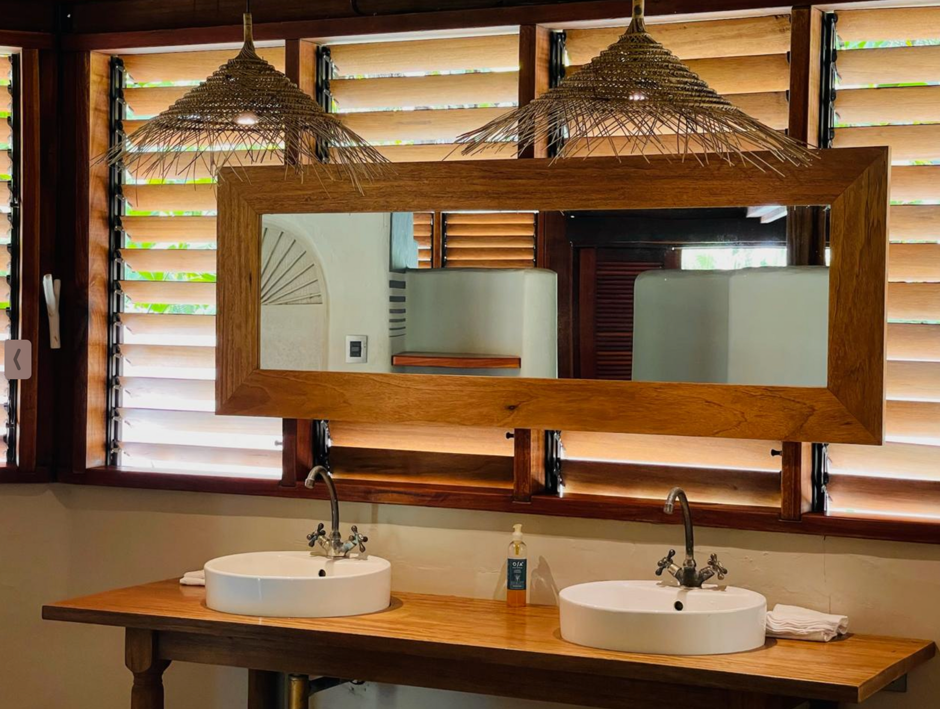 Wooden bathroom vanity with two sinks, large framed mirror, and woven pendant lights against a windowed wall