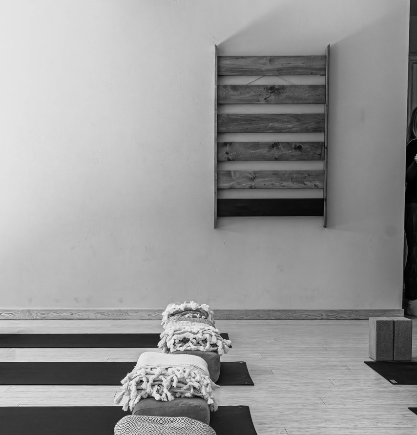 Person with tattooed back raises hands in a yoga studio with mirrors and large windows.