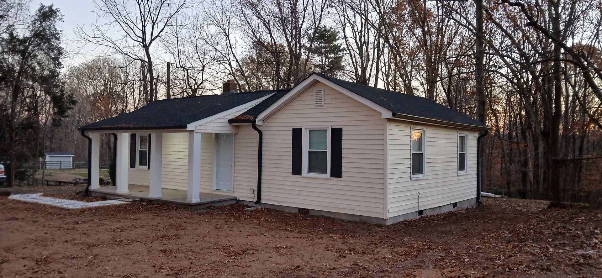 Beige house with a black roof and black shutters, set in a brown, wooded area.
