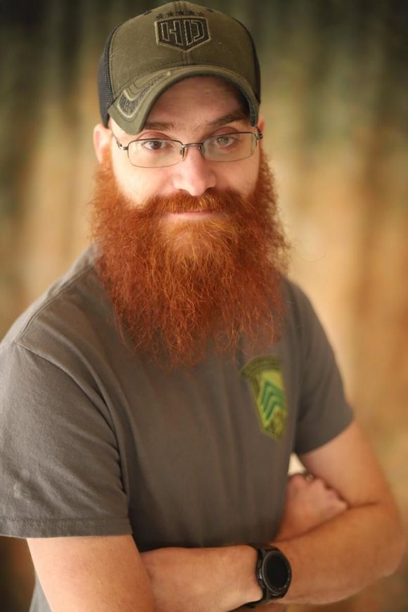 Portrait of a bearded man in a green cap and gray shirt with arms crossed, smiling at the camera.