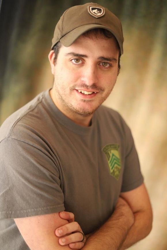Man in brown cap and gray T-shirt smiling with arms crossed against a blurred background