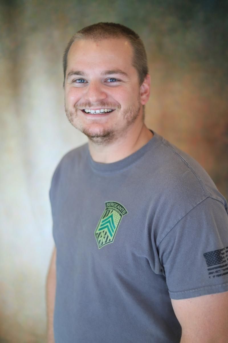 Smiling person in a gray T-shirt with a green logo, standing against a blurred studio backdrop