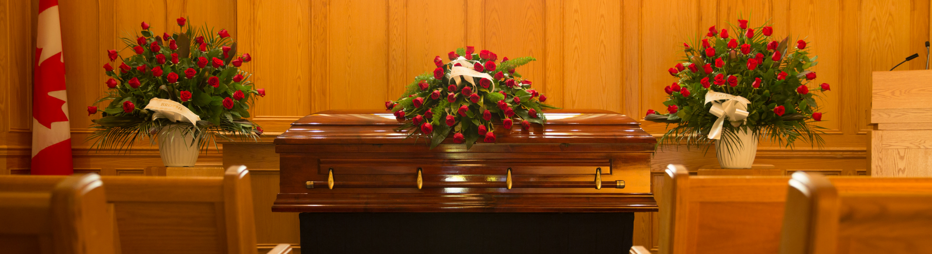 Coffin on a wooden casket table, framed by red and white floral arrangements in a funeral chapel