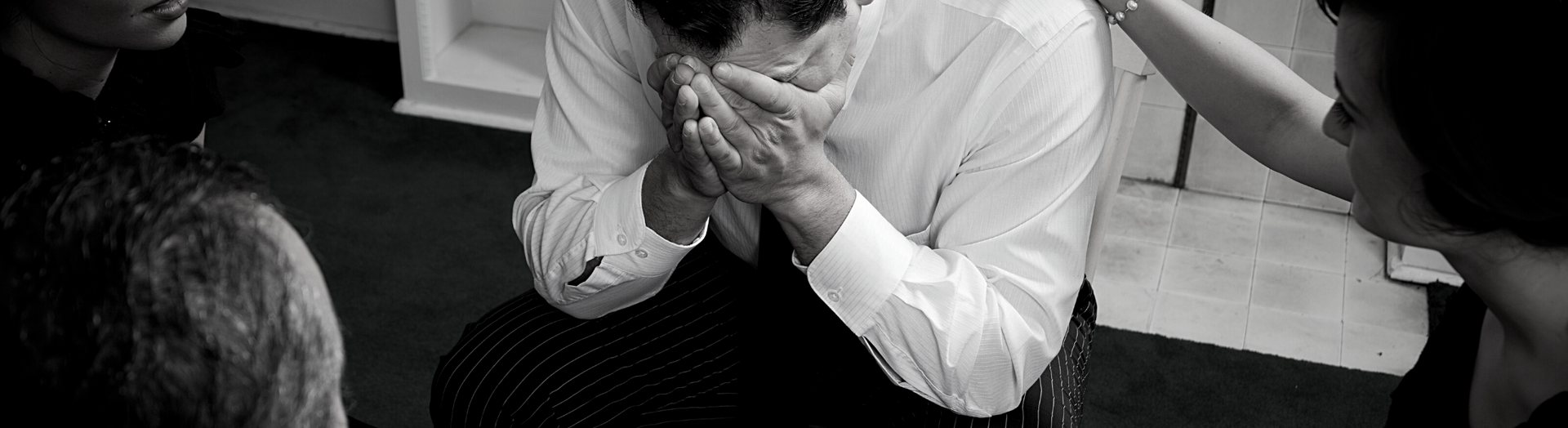 Man in a white shirt covers his face while seated, with another person beside him in a black-and-white scene