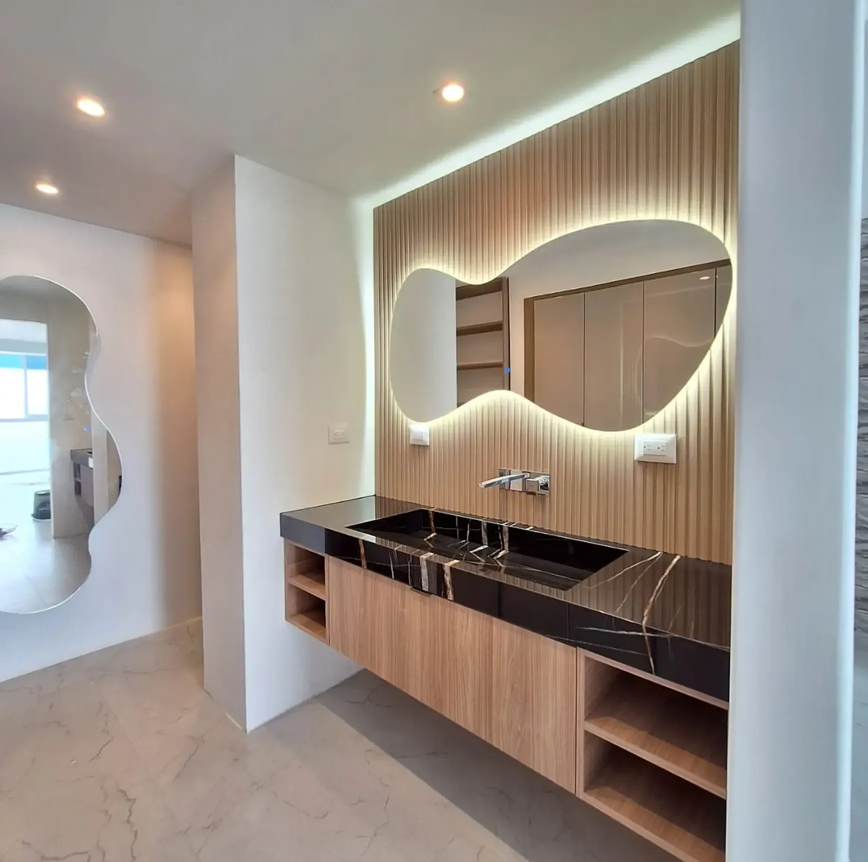 Tocador de baño moderno con encimera negra, revestimiento de madera y espejo redondo retroiluminado en un interior luminoso.