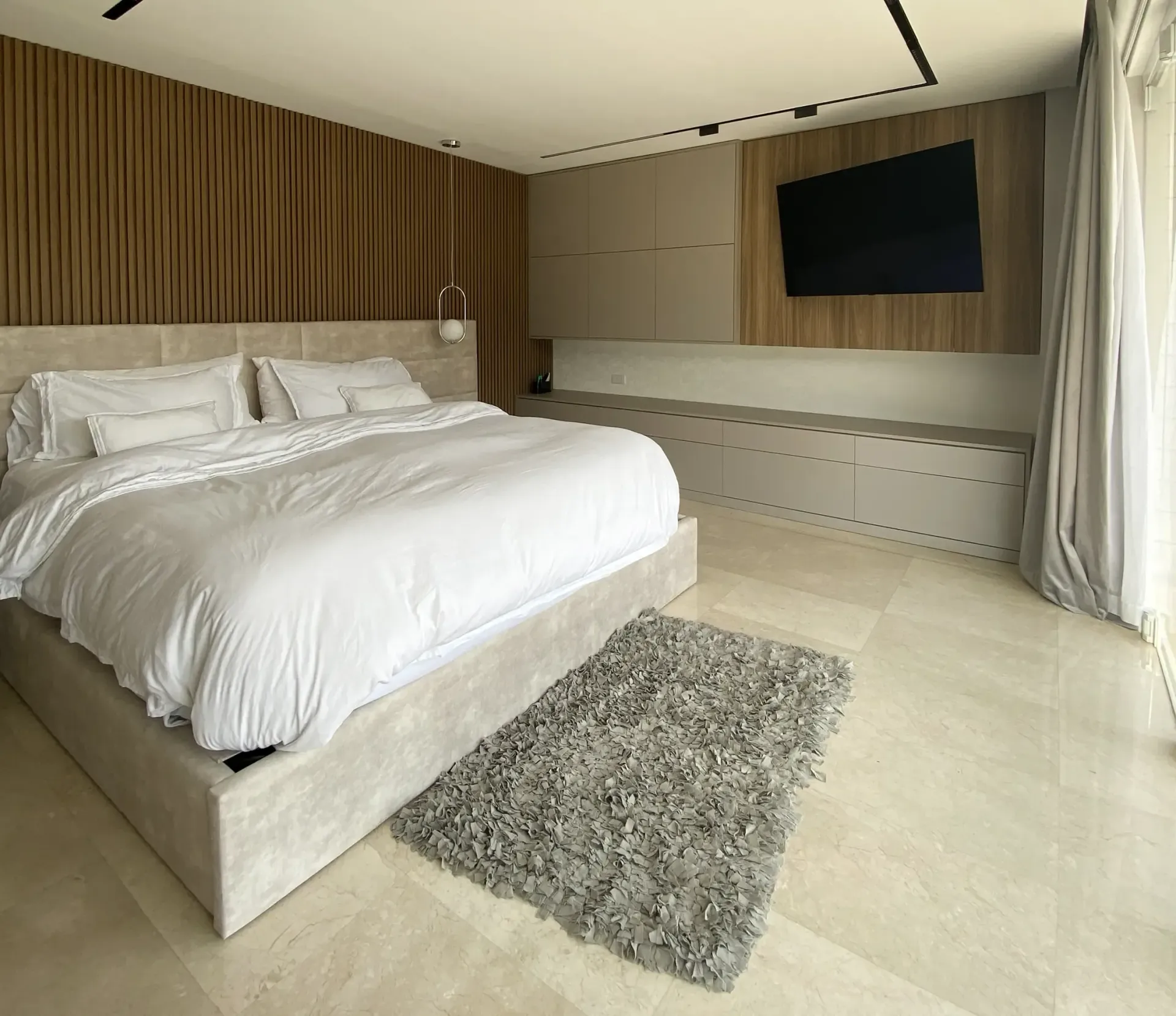 Dormitorio moderno con una cama grande, pared revestida de madera, televisor montado en la pared y alfombra de pelo largo junto a cortinas transparentes.