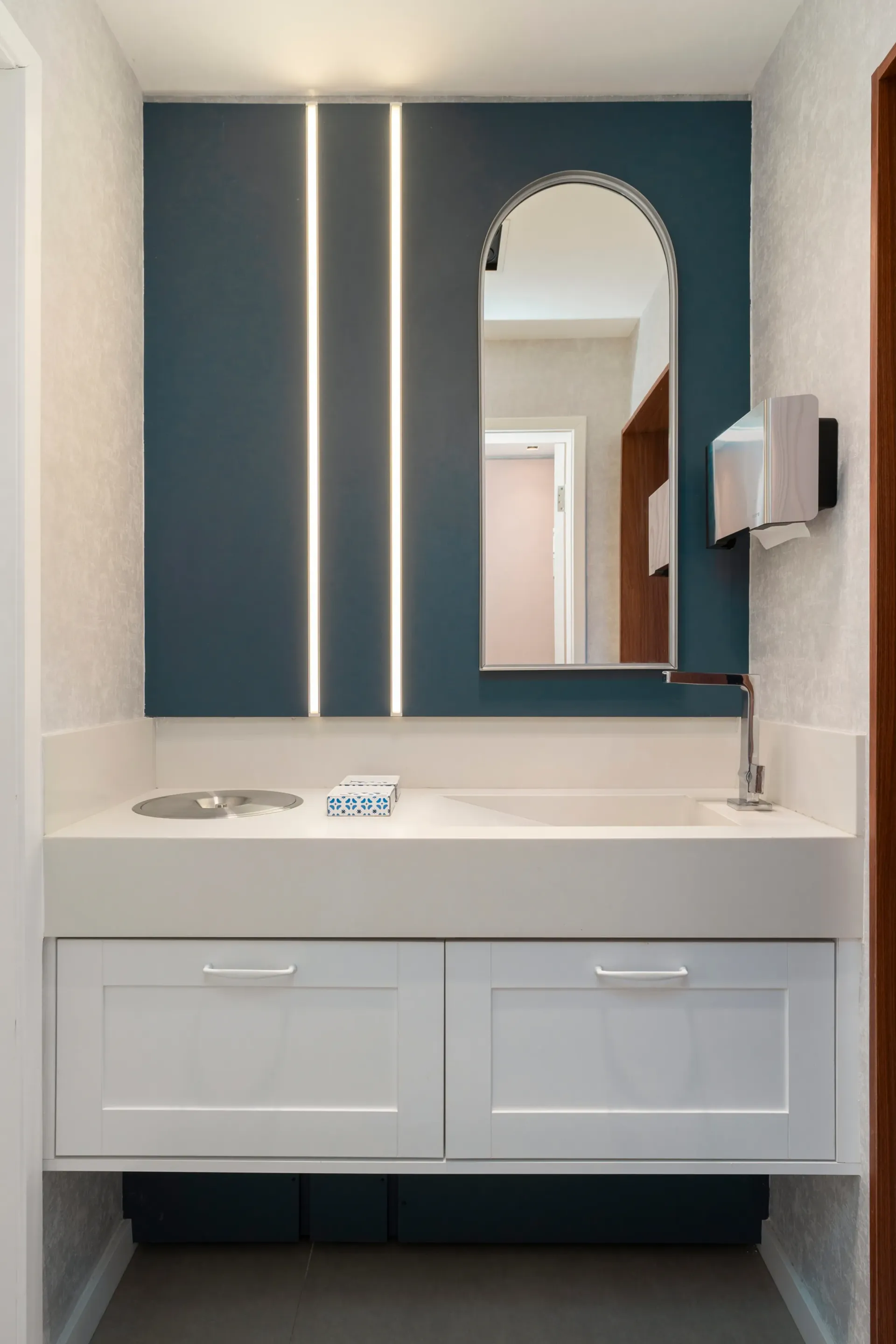Tocador de baño moderno con lavabo blanco, pared decorativa azul y espejo arqueado con iluminación.