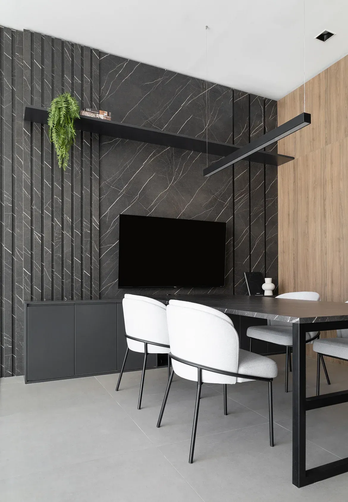 Rincón de comedor moderno con pared decorativa negra, televisor montado en la pared, sillas blancas y una planta colgante.