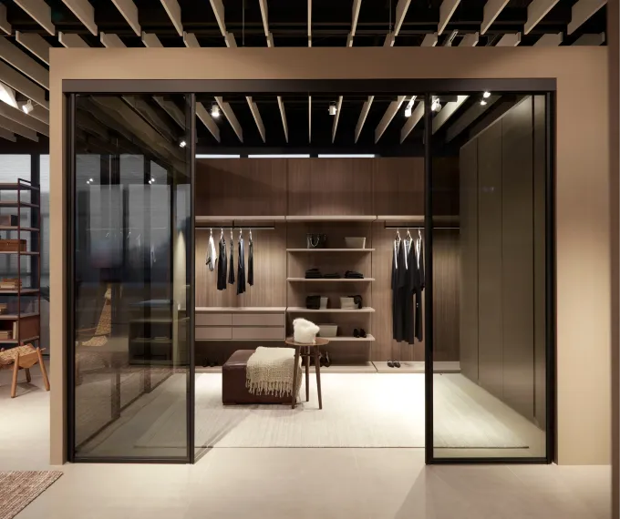 Vestidor moderno con puertas de cristal, estantes de madera, perchero y banco central.