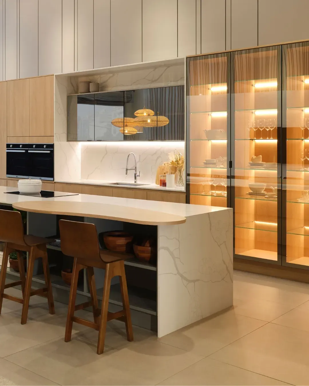 Cocina moderna con gabinetes de madera, isla de mármol, taburetes de bar y refrigerador con puerta de vidrio iluminado en el interior.