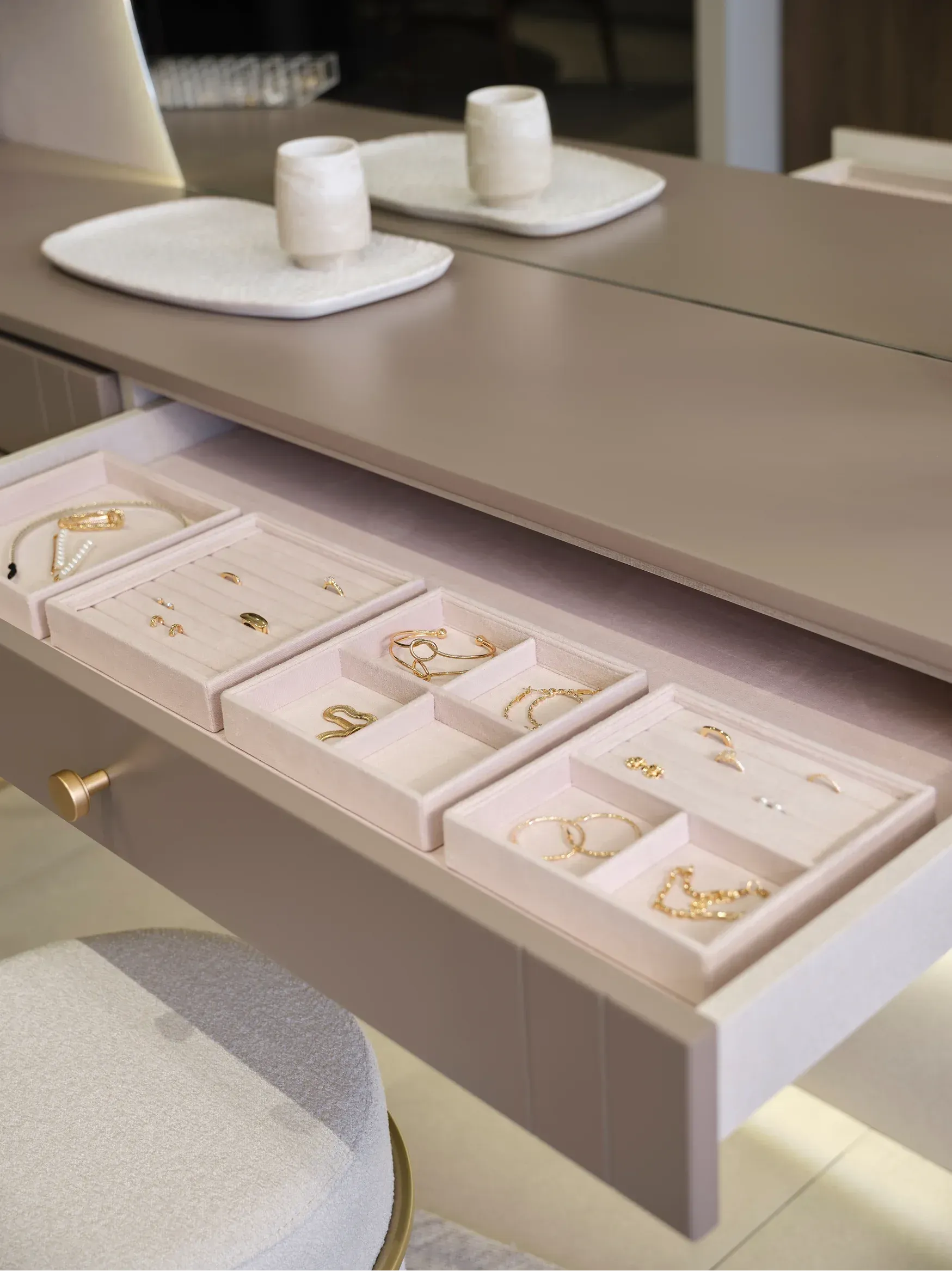 Cajón de joyería abierto debajo de un tocador beige con bandejas para anillos y dos frascos blancos encima.