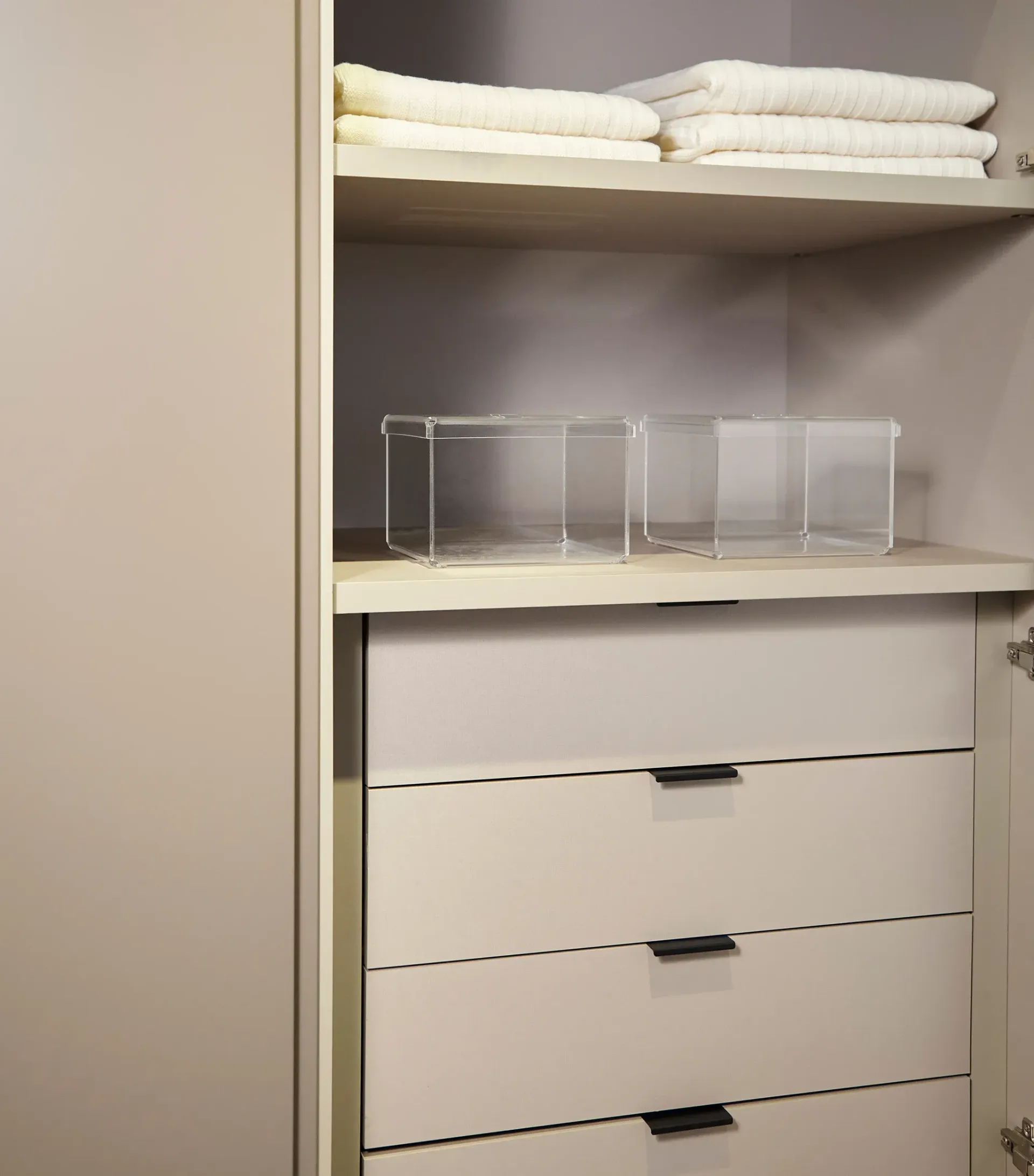 Closet abierto con toallas blancas dobladas, cajas de almacenamiento transparentes y cajones blancos.