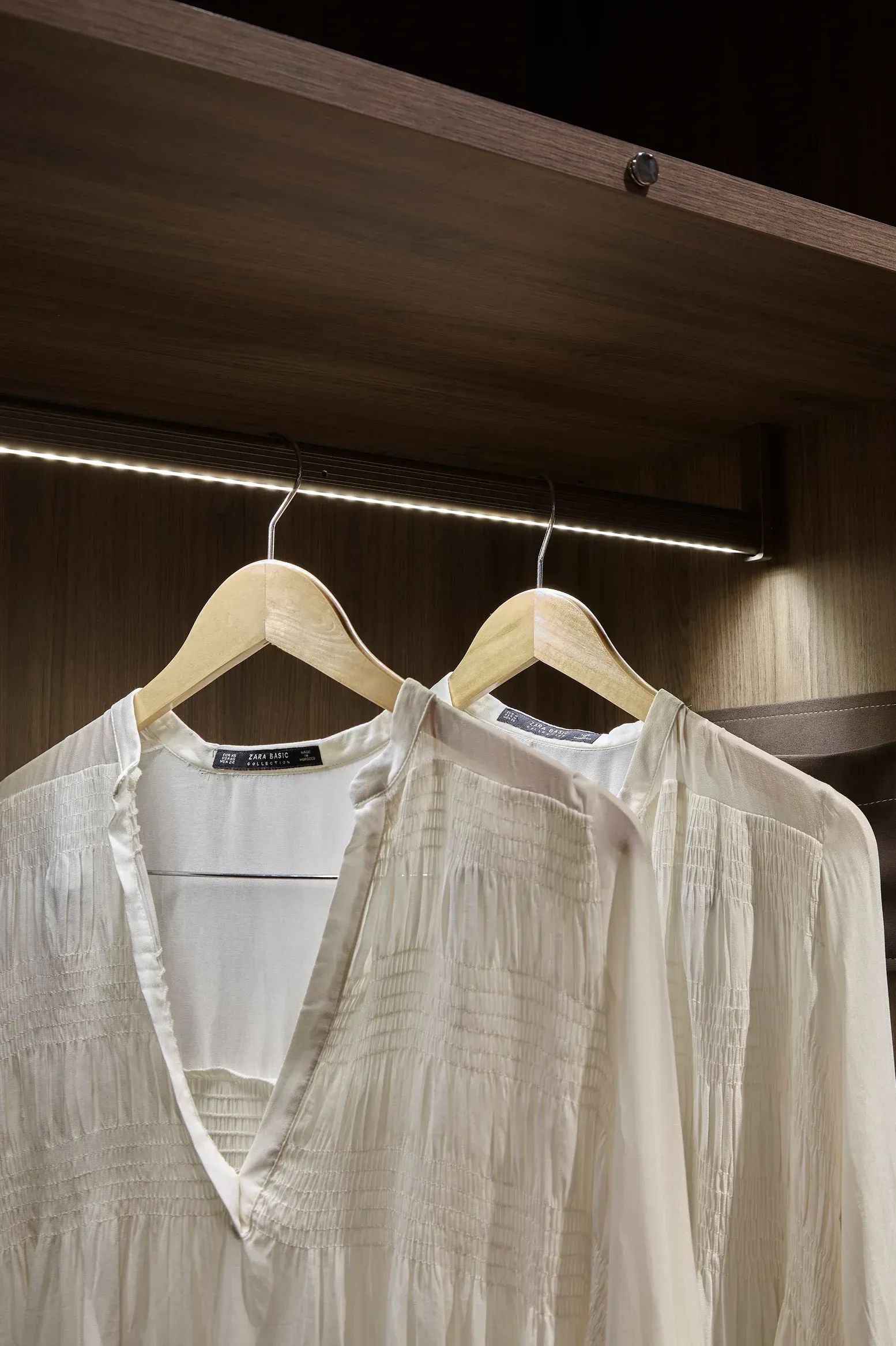 Blusas blancas en perchas en un armario de madera con iluminación en los estantes.