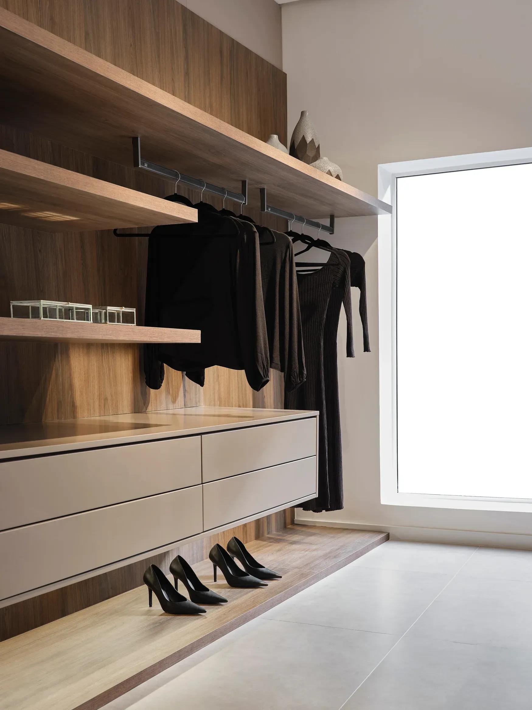Vestier moderno con estantes de madera, perchero negro, cajones y espacio para zapatos junto a una ventana luminosa.