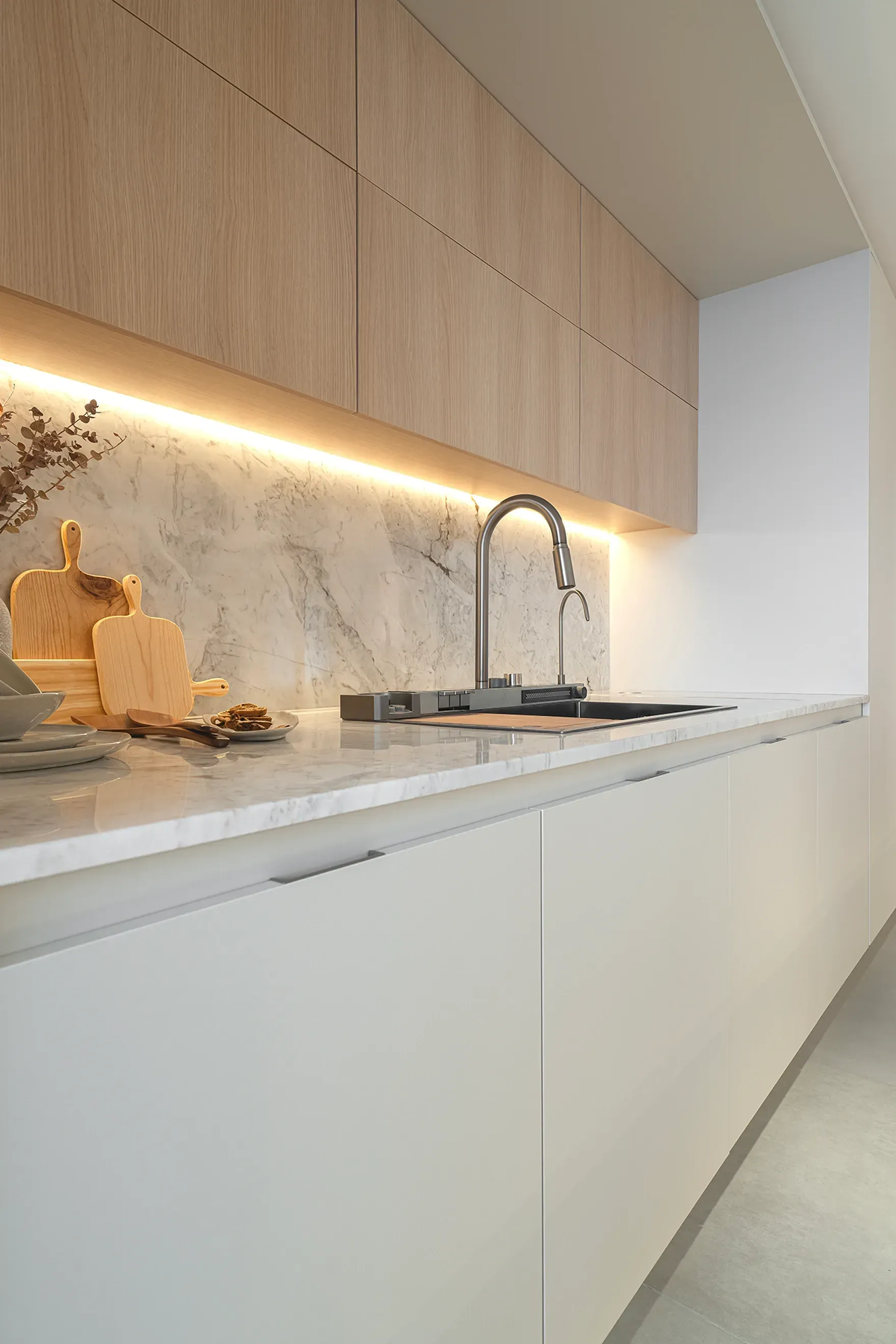 Cocina moderna minimalista con gabinetes blancos, gabinetes superiores de madera, protector contra salpicaduras de mármol y encimera iluminada por grifo.