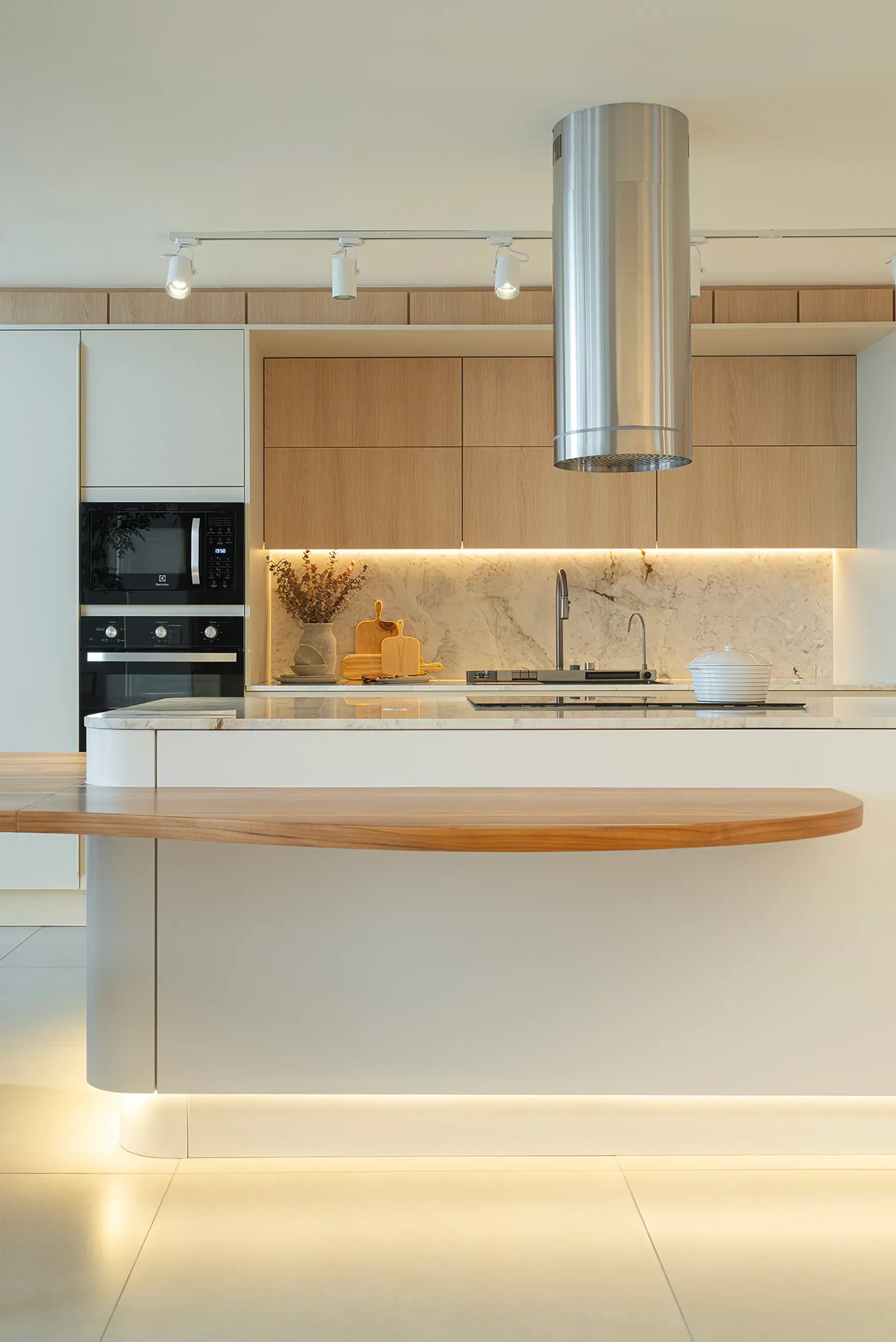 Cocina moderna minimalista con isla blanca, detalles en madera, iluminación bajo los gabinetes y campana extractora de acero inoxidable.