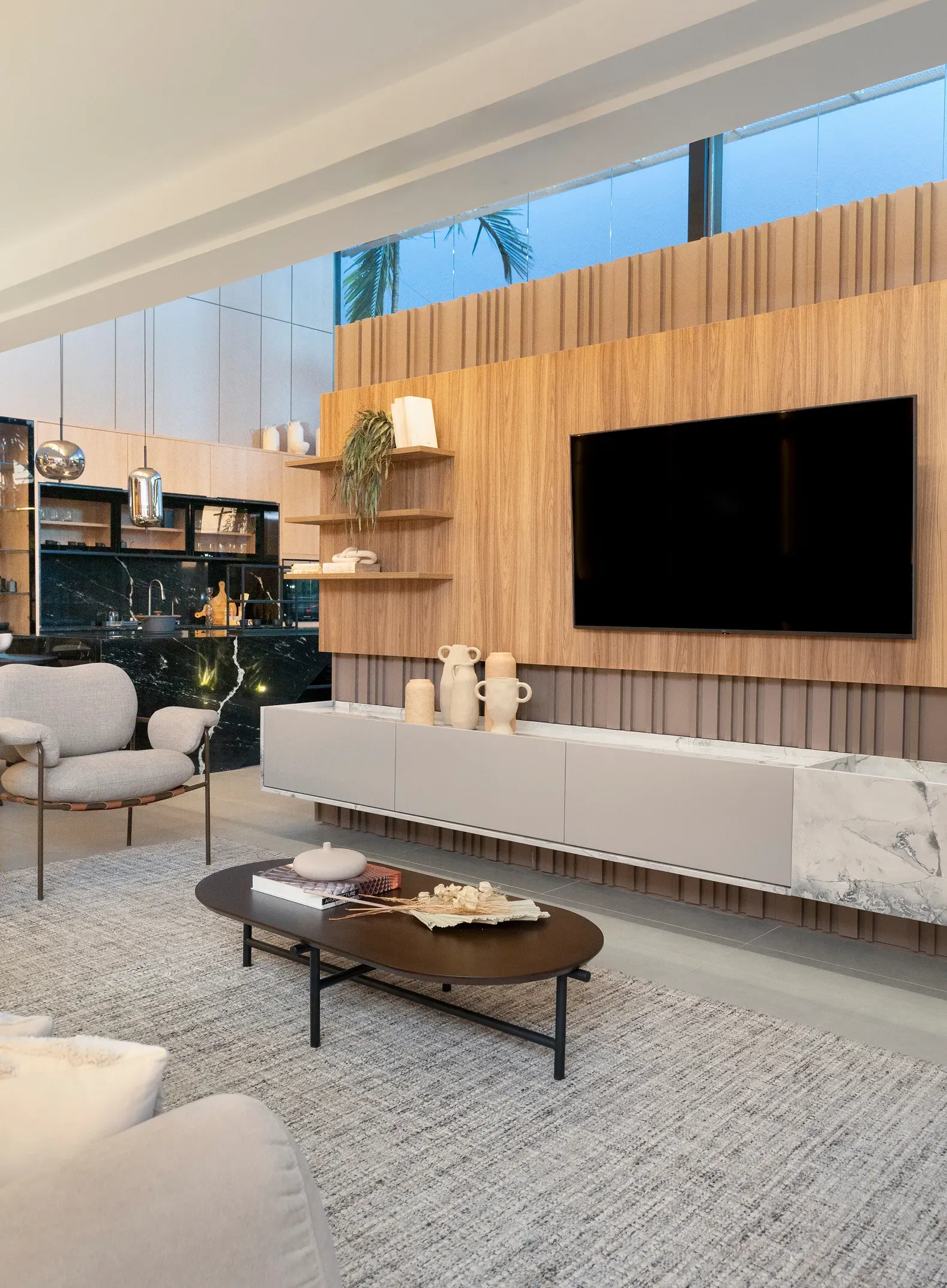 Sala de estar moderna con pared de madera para el televisor, televisor negro, sofá blanco, mesa de centro y cocina abierta al fondo.