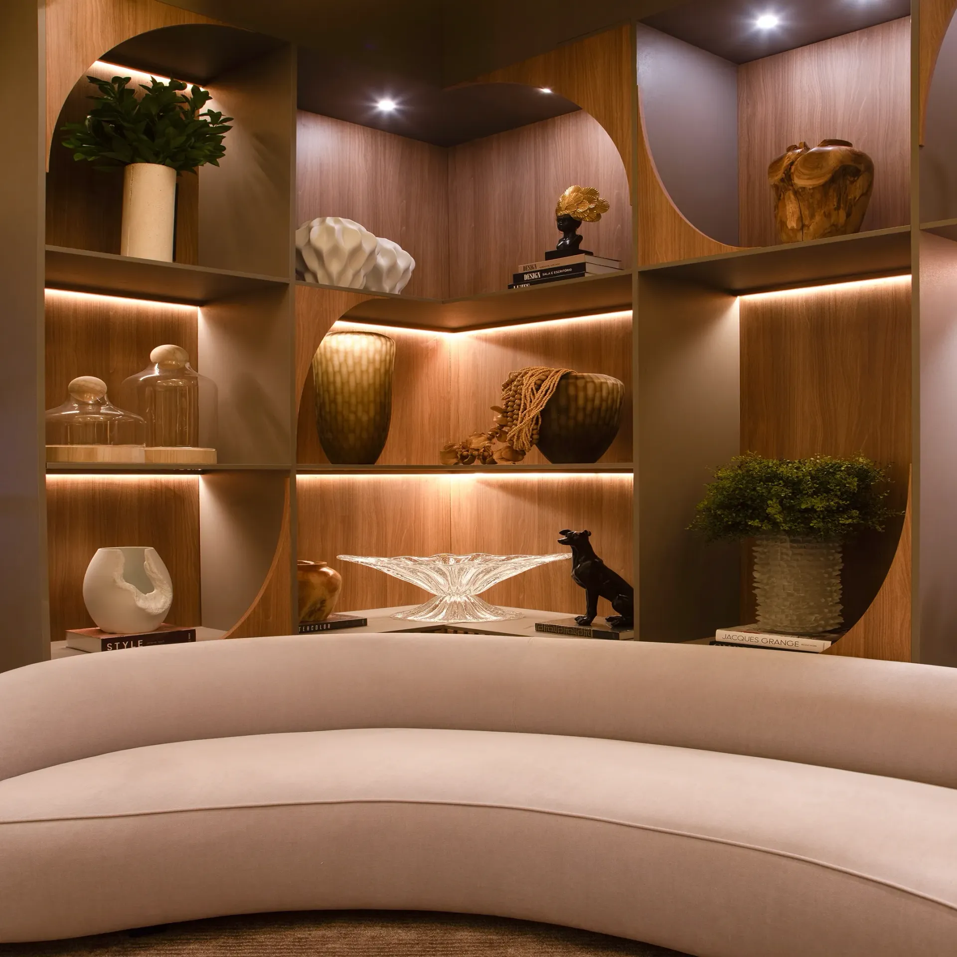 Sofá curvo de color beige frente a estantes de madera iluminados con esculturas decorativas y plantas.