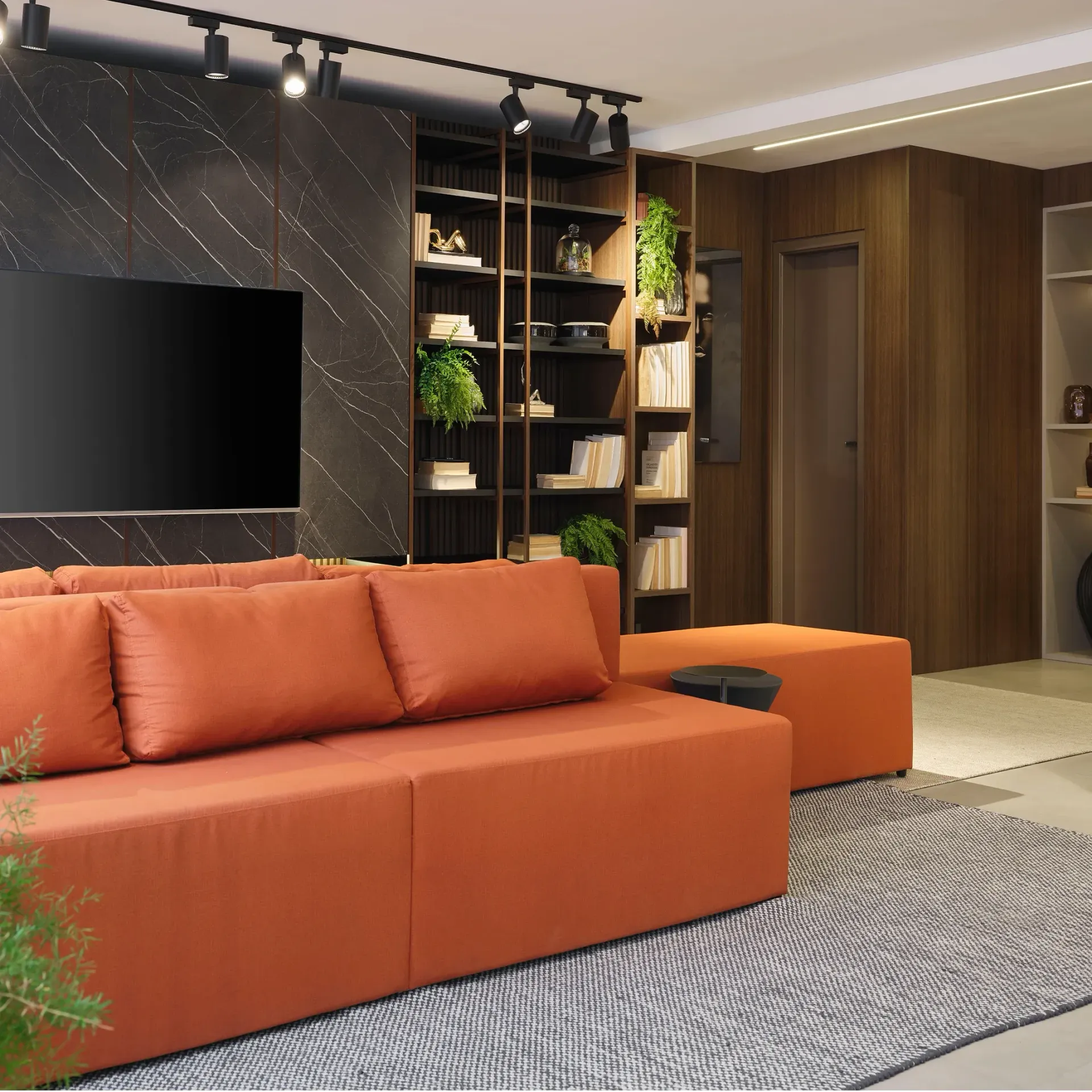 Sala de estar moderna con sofá seccional naranja, televisor montado en la pared, estanterías de madera y alfombra texturizada.
