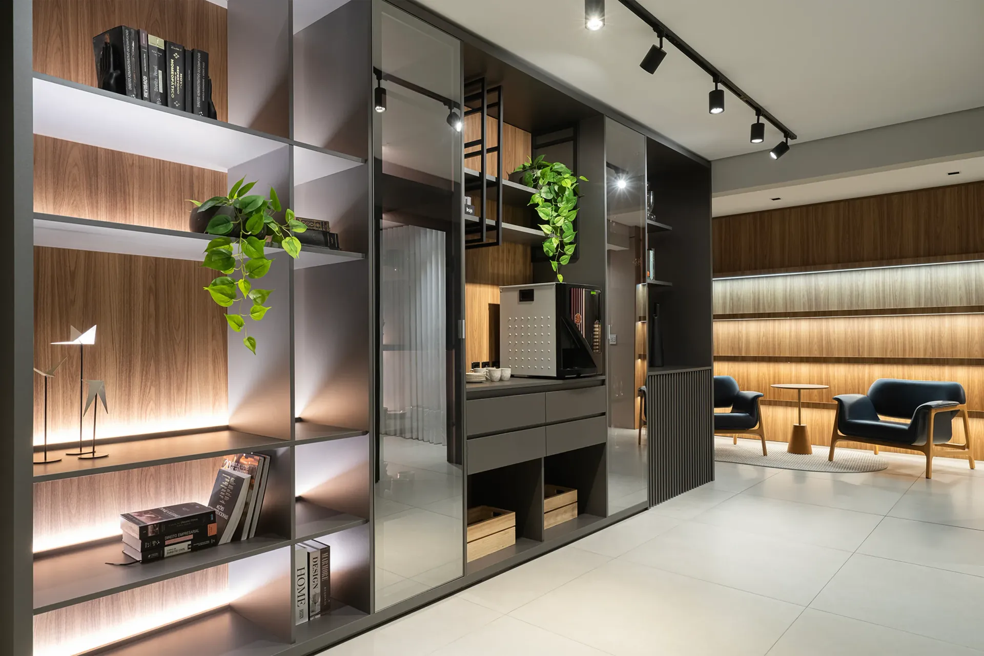 Sala de estar moderna con mamparas de cristal, estanterías de madera cálida, plantas colgantes y sillas bajo iluminación de riel.