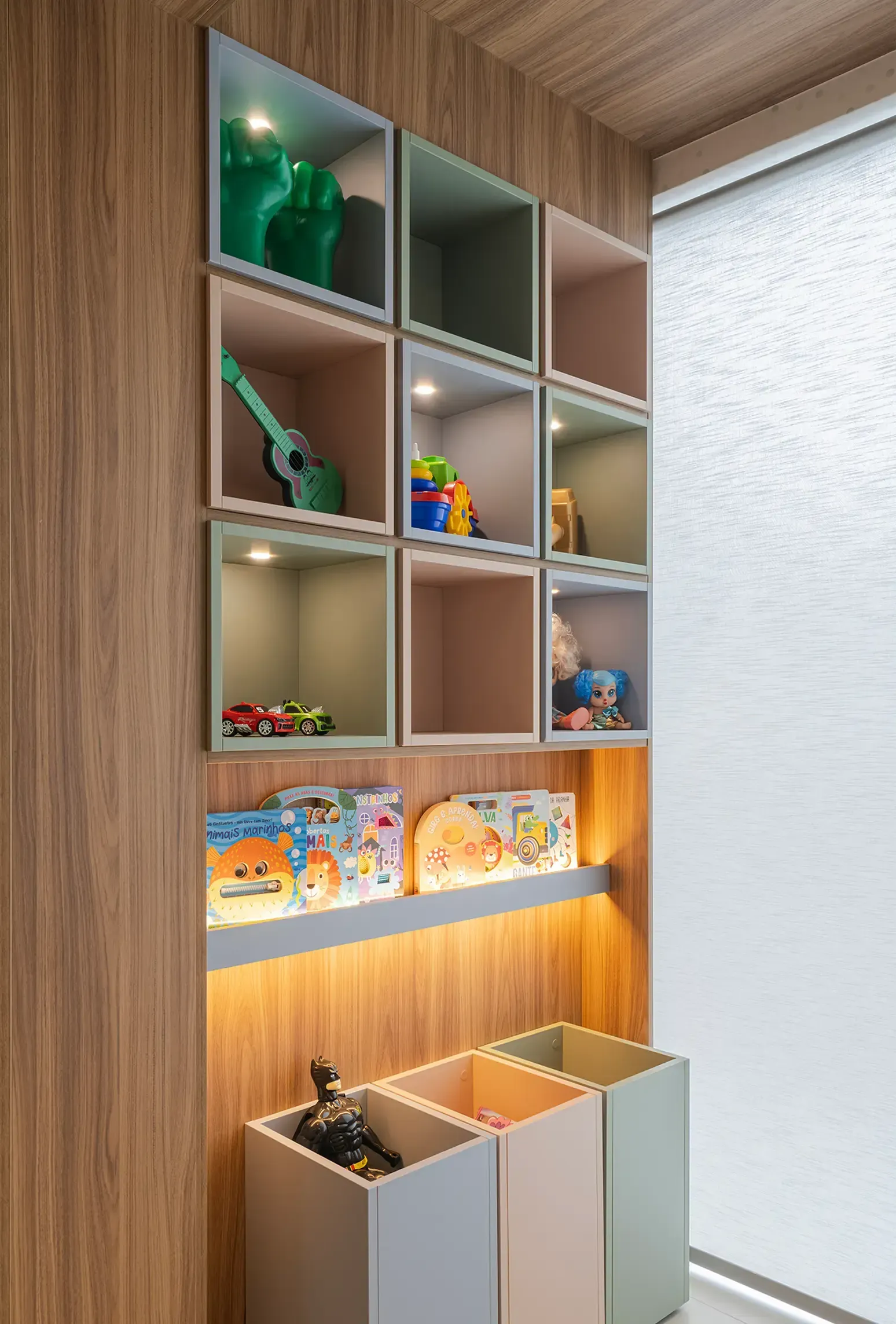 Estanterías de madera en la pared con juguetes y libros coloridos sobre tres cajas de almacenamiento en un rincón luminoso.
